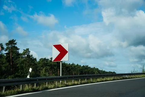 Red and white chevron direction sign beside a curving road with green trees Stock Photos