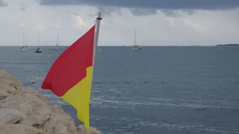 A red and yellow warning flag on a rocky point, sailboats moored in a bay under Stock Footage 344818561
