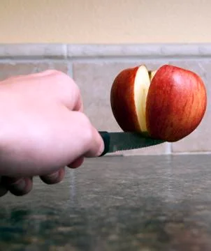 Red apple being cut in half with a sharp knife being held up by one single to Stock Photos