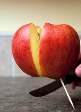 Red apple being cut in half with a sharp knife being held up by one single to Stock Photos