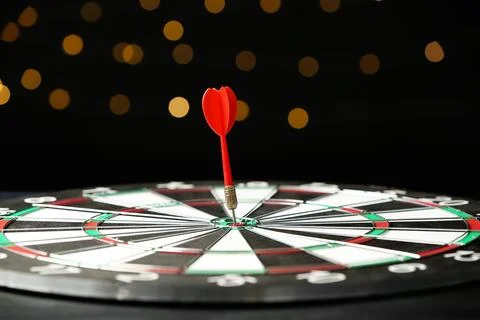 Red arrow hitting target on dart board against blurred lights Stock Photos