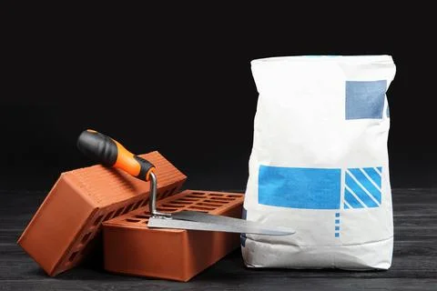 Red bricks, bag of cement powder and putty knife on dark wooden table. Buil.. Stock Photos