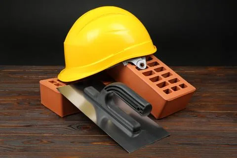Red bricks, putty knife and yellow hard hat on wooden table. Building materia Stock Photos