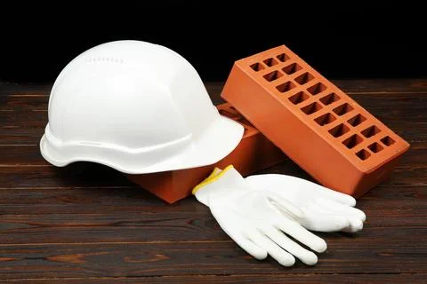 Red bricks, white hard hat and rubber gloves on wooden table. Building materi Stock Photos