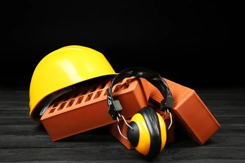 Red bricks, yellow hard hat and earmuffs on dark wooden table. Building mat.. Stock Photos