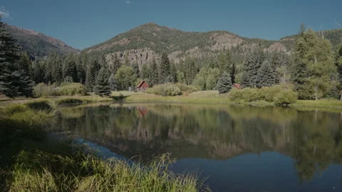 Red cabin reflected in a calm mountain pond below rocky peaks Colorado Stock-Footage 348023725