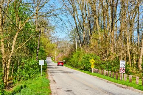 Red car driving down side road in midwest late spring with budding leaves Fotos de archivo
