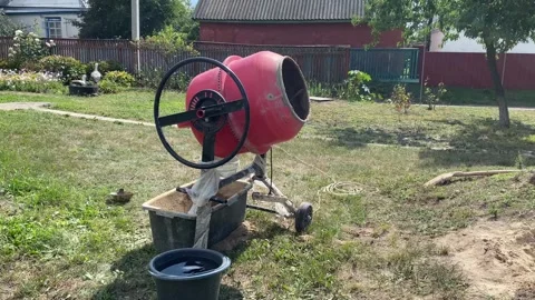 Red cement mixer rotating in yard with black water bucket and clay container Stock Footage 316539392