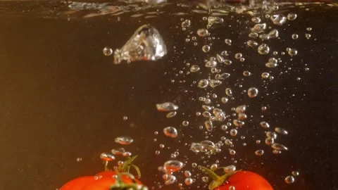 Red cherry tomatoes falling into water with air bubbles. Black background. Slow Stock Footage