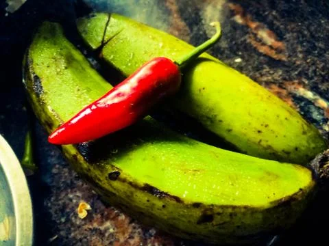 A Red chilli placed above two green bananas on a kitchen table top. Stock Photos