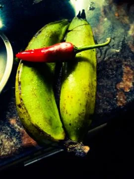 A Red chilli placed above two green bananas placed on a kitchen table top. Stock Photos