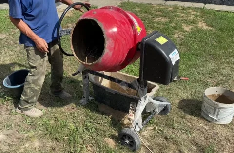 Red concrete mixer working as man pours water from black bucket Stock Footage 316539402