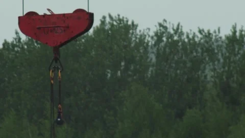 Red construction crane hook against blurred green trees and sky, industrial Stock Footage 329213745