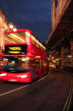 Red double-decker bus speeding through central London streets at dusk. Stock Photos