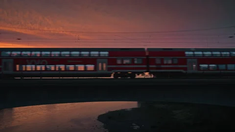 Red double-decker train crossing a bridge at sunset with illuminated windows Stock Footage 315179750