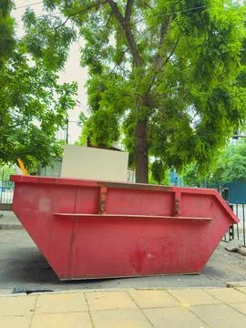 Red dumpster located on a city street surrounded by green trees and buildin.. Stock Photos