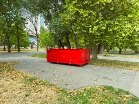 A red dumpster is placed on a concrete path in a park. Trees surround the are Stock Photos