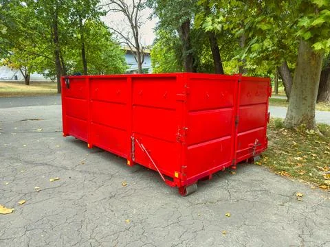 A red dumpster sits on the cracked ground in a park. Tall trees provide shade Stock Photos