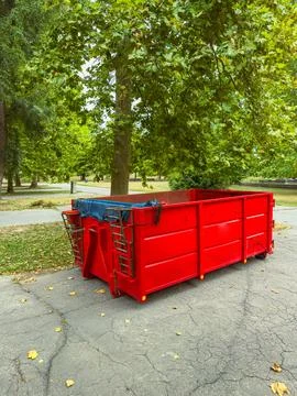 A red dumpster sits on a paved surface in a park. Trees with green leaves sur Foto stock