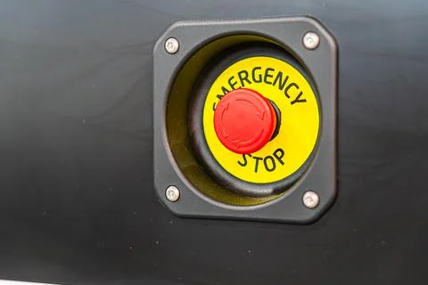 A red emergency stop button, label visible, set against a dark, indistinct ba Stock Photos