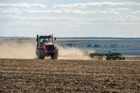 A red farm tractor in a cloud of dust cultivates the soil in the field with a Stock Photos