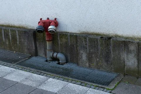Red fire hydrant positioned alongside concrete wall with attached pipe Stock Photos