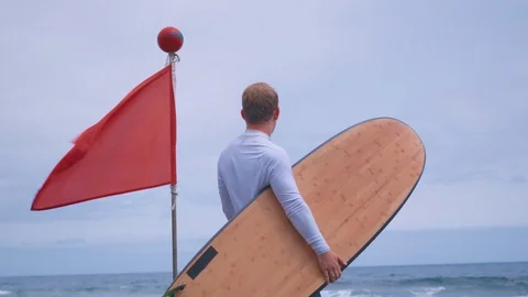 A red flag means danger and a surfer with a surfboard stands nearby and looks at Stock-Footage 101854203