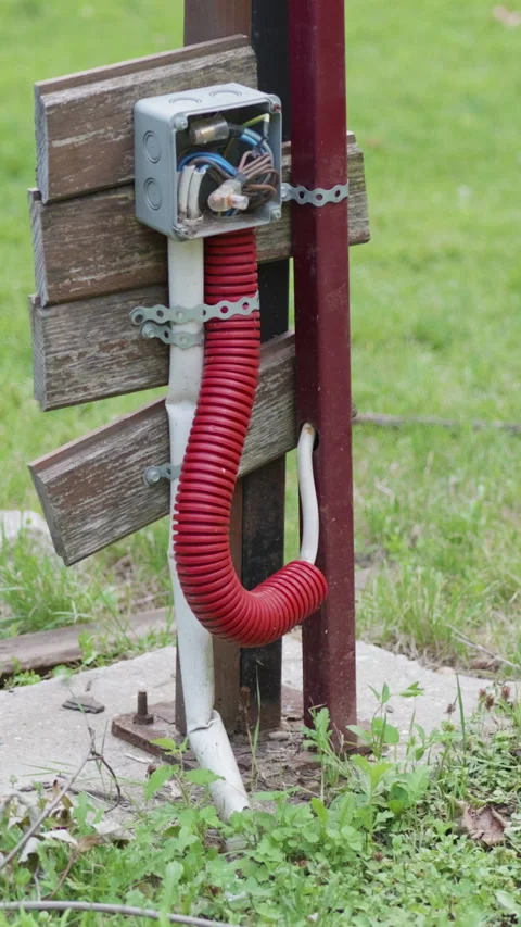 Red flexible hose connected to electrical box on a wooden post in a grassy area Stock Footage 341439830