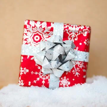 Red gift box with a silver bow on a snowdrift on a kraft background.. Stock Photos