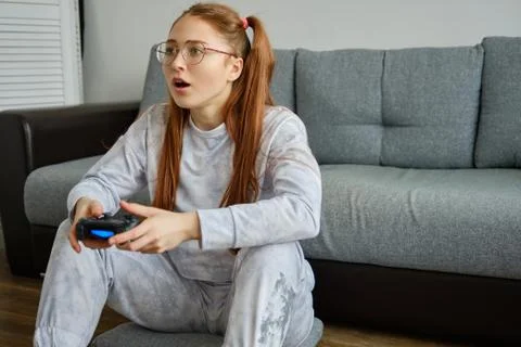 Red girl with tails in glasses sits on the floor and enthusiastically plays Stock Photos