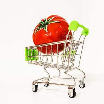 A red glossy tomato lies in a tiny metal supermarket cart on an isolated Stock Photos