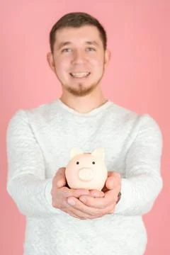 A red-haired bearded man in a white sweater holds out a piggy bank standing o Stock Photos