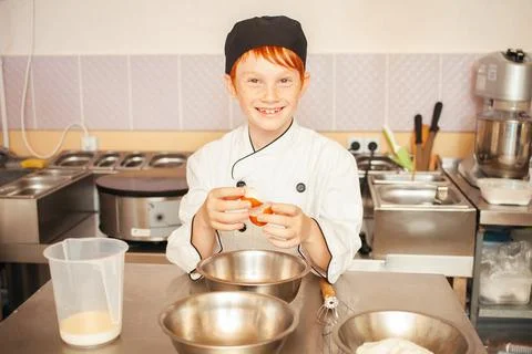 Red-haired boy in chef's costume cooks pancakes in kitchen, kneads dough Stock Photos