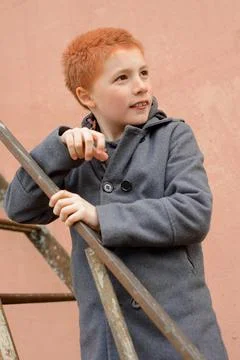 Red-haired boy in a grey coat climbed the rusty metal ladder to Stock Photos
