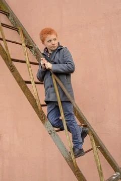 Red-haired boy in a grey coat climbed the rusty metal ladder to the wall and  Stock Photos