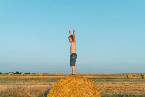 A red-haired boy stands with hands up on top of a straw bale on a wheat field Stock Photos