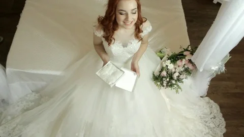 Red-haired bride in a white lavish dress sits on the bed and enjoys the gift Stock Footage 82473902
