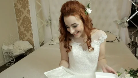 Red-haired bride in a white lush dress sits on the bed and opens a white box Stock Footage 82469699