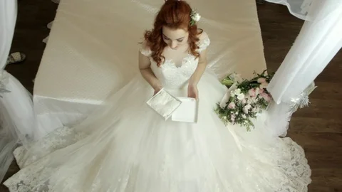 Red-haired bride in a white luxurious dress sits on the bed and opens a gift Stock Footage 83246870