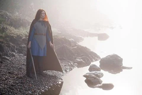 Red-haired girl in armor with sword and raincoat on river bank Stock Photos
