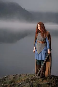 Red-haired girl in armor with sword and raincoat on river bank Stock Photos