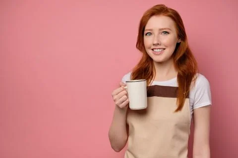 Red-haired girl in a beige apron and a T-shirt stands on a pink background and Stock Photos