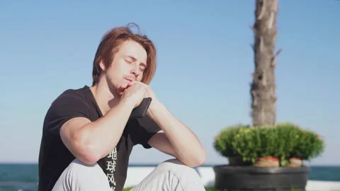 Red-haired guy in a black T-shirt sits on the background of sea and blue sky 動画素材 163411890