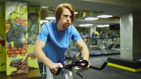 Red-haired guy in a blue t-shirt rides a bicycle in the gym. Stock Footage 163458689