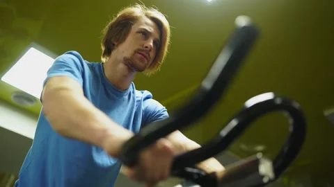 Red-haired guy in a blue t-shirt rides a bicycle in the gym. close-up of a hand Vidéo 163458898