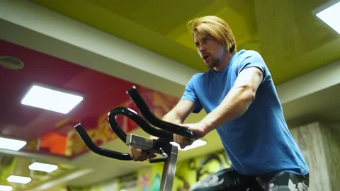 Red-haired guy in a blue t-shirt rides a bicycle in gym.on a yellow background Vidéo 163459904