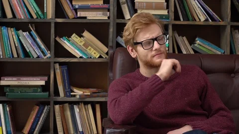 Red-haired man sitting in library taking off glasses Stock Footage 121589599