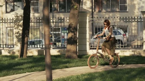 Red haired woman riding bicycle in city park on sunny day Stock Footage