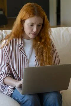 Red-haired woman on a sofa using a laptop for remote work and online study,.. Stock Photos