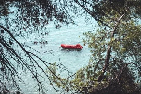 Red inflatable boat moored in clear turquoise sea framed by coastal pine tr.. Stock Photos